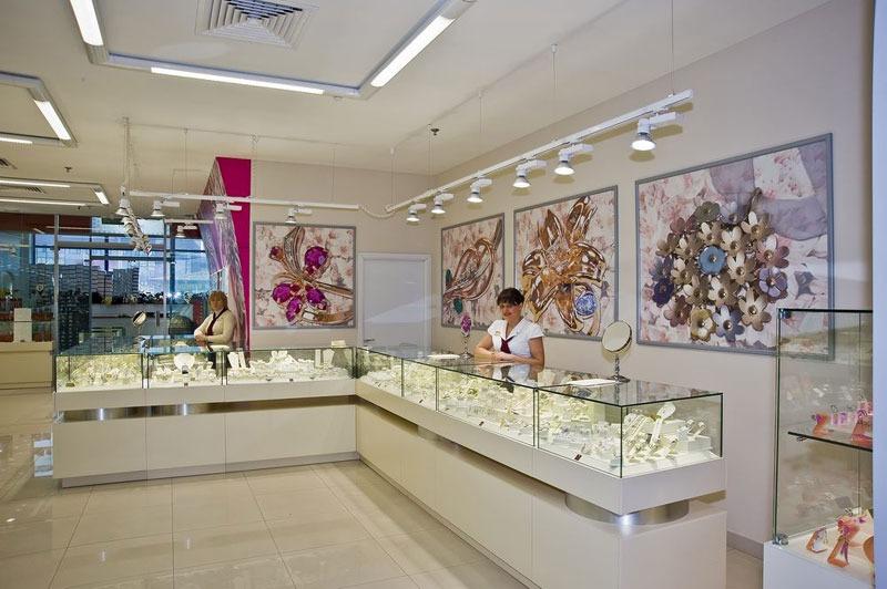 Bright jewelry salon with white glass-top counters, island vitrines and a tall display cabinet.