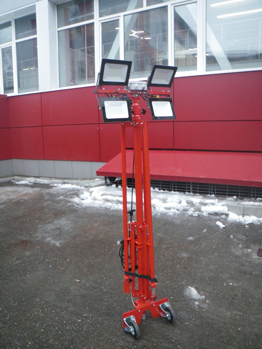 Red wheeled lighting mast with four LED floodlights on a snowy outdoor surface.