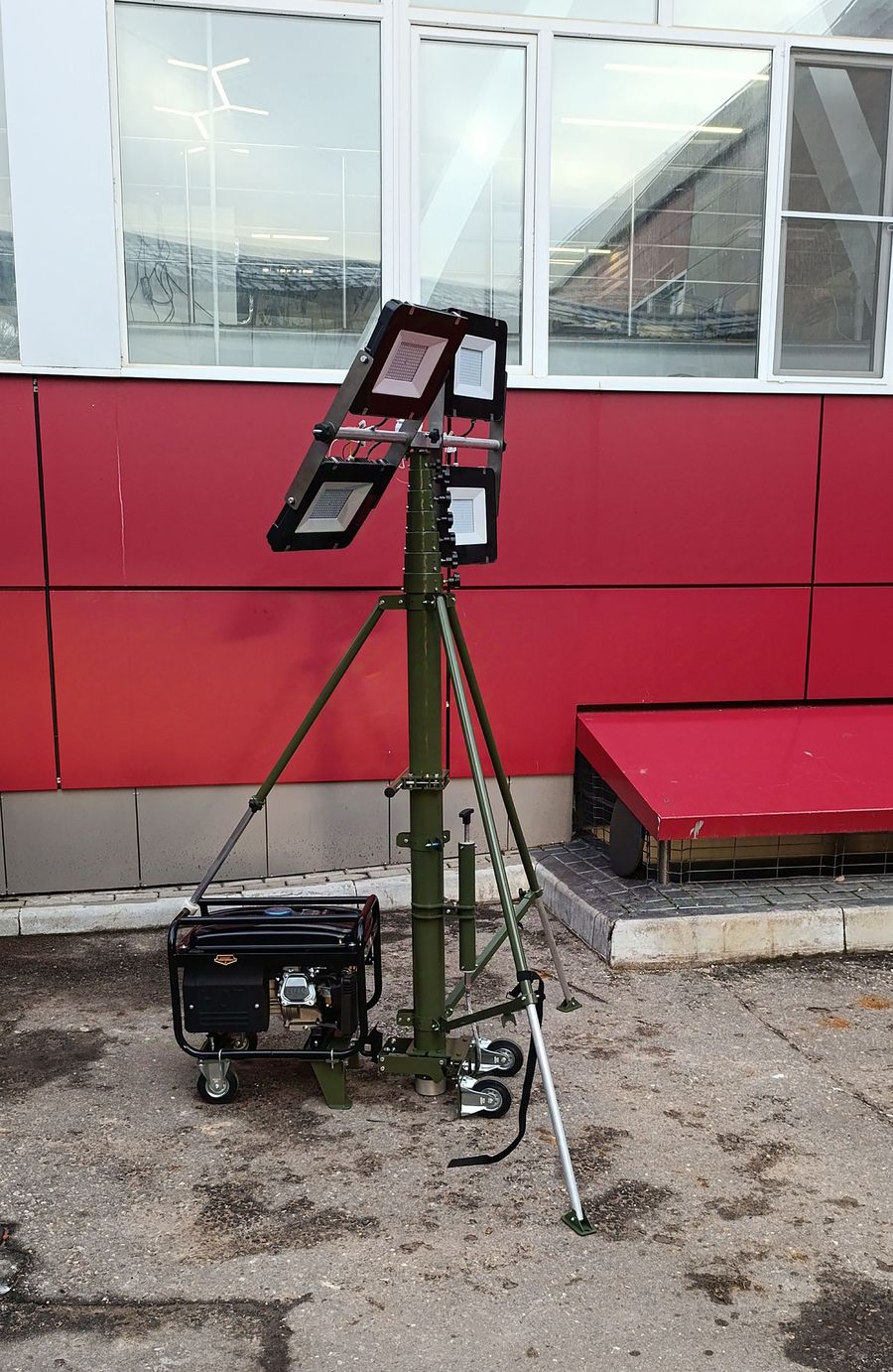 Olive-green telescopic lighting mast on a wheeled tripod with four LED floodlights and a portable generator.