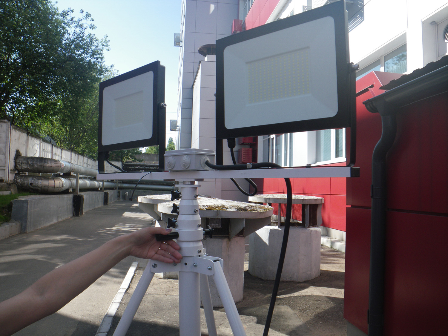 White portable lighting mast with two LED floodlights on a tripod, outdoors beside a building.